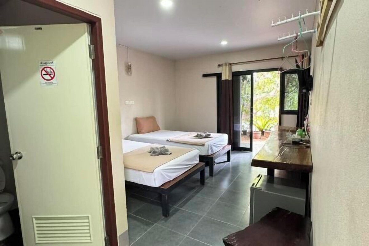 A room with two beds, a small table, a bench, a door with a no-smoking sign, and a view of greenery outside.