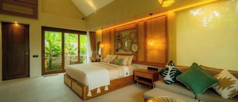 A bedroom with a large bed, a sofa, and a dining table. There are wooden doors and a window with a view of greenery.