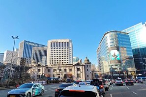A busy urban street with modern buildings, including a prominent glass-fronted structure. - QINGHE HOME (Seoul)