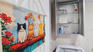 A bathroom with a wall-mounted shelf containing toiletries, a painting of cats, and a Samsung washing machine. - QINGHE HOME (Seoul)