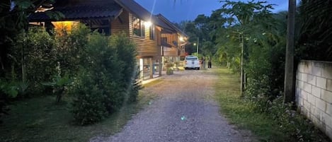 Un camino de grava conduce a una casa de madera con luz exterior, rodeada de follaje.