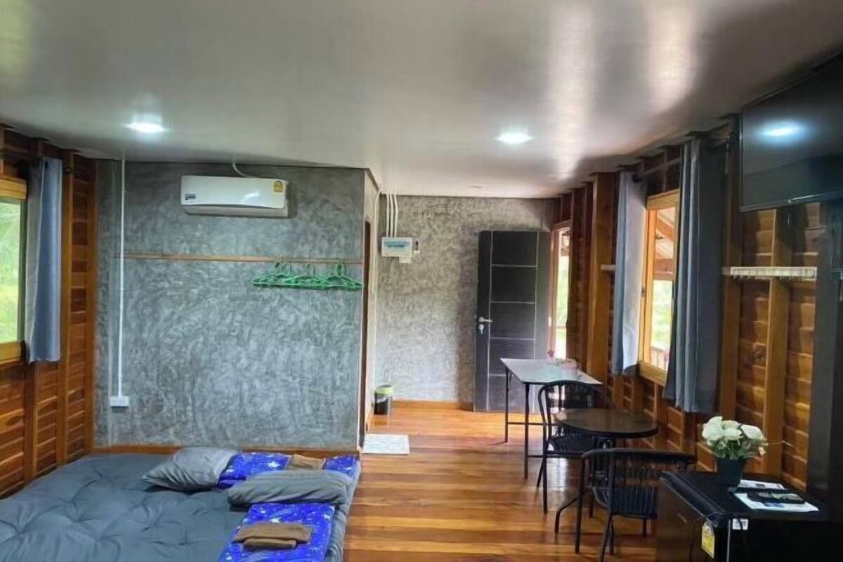A compact room with a bed, a small table, and a chair. There is an air conditioning unit on the wall, and the room has wooden paneling on the walls.
