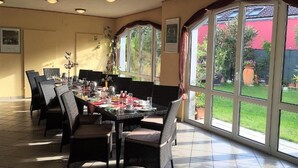 A dining area with a long table, wicker chairs, and large windows with a view of greenery. - Pension Haus Marianne (Nußdorf ob der Traisen)