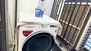 Una lavadora de carga frontal con tapa blanca, una bolsa para detergente encima y una reja metálica al fondo.