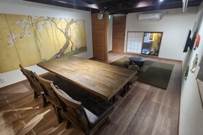 Ruokailutila, jossa on puinen pöytä ja tuolit, tatamimatto ja perinteinen japanilainen maalaus seinällä.