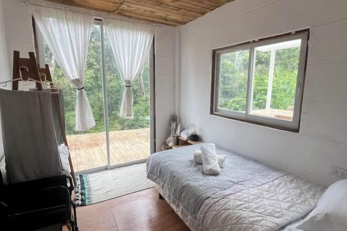 A bedroom with a bed, a window, a curtain, and a view of greenery outside.