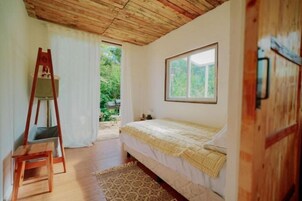 A bedroom with a wooden ceiling, a bed with a white and yellow bedspread, a window, and a wooden ladder against the wall.