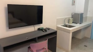 Une chambre moderne dotée d’un téléviseur à écran plat fixé au mur, d’un petit bureau avec une chaise et d’un coin cuisine intégré comprenant un évier et un four à micro-ondes.