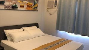 Un lit bien fait, avec une literie blanche et une tête de lit décorative, un climatiseur fixé au mur et une fenêtre ornée de rideaux légers.