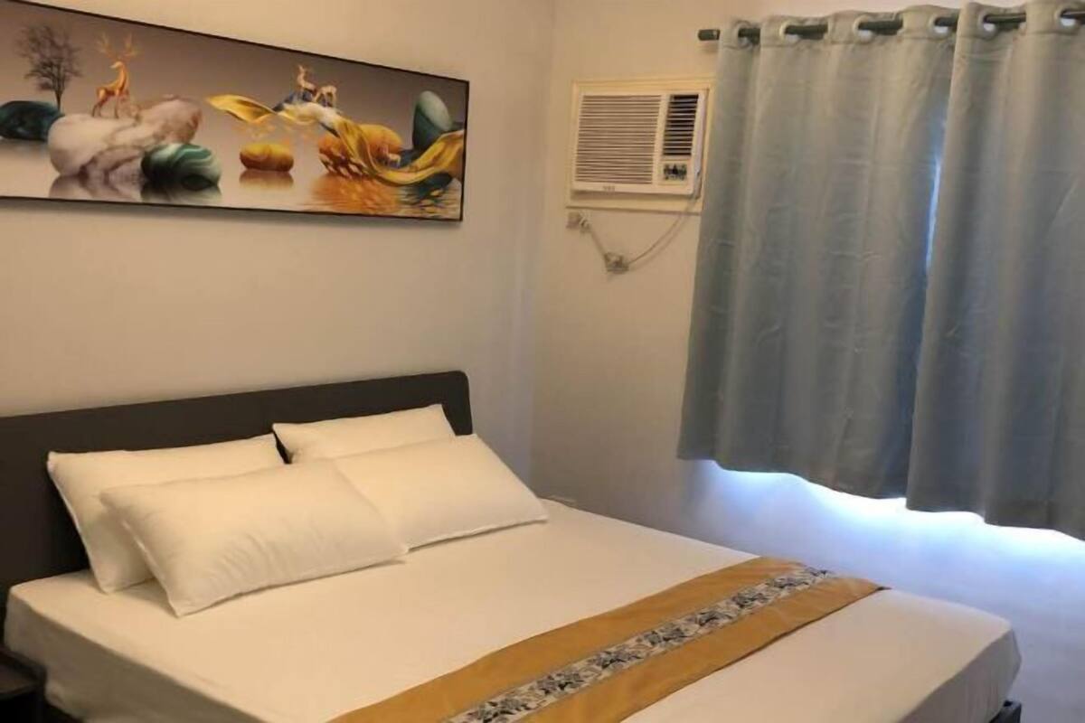 Ένα στρωμένο κρεβάτι με λευκά σεντόνια και ένα διακοσμητικό προσκέφαλο, μια μονάδα κλιματισμού στον τοίχο και ένα παράθυρο με ελαφριά κουρτίνες.
