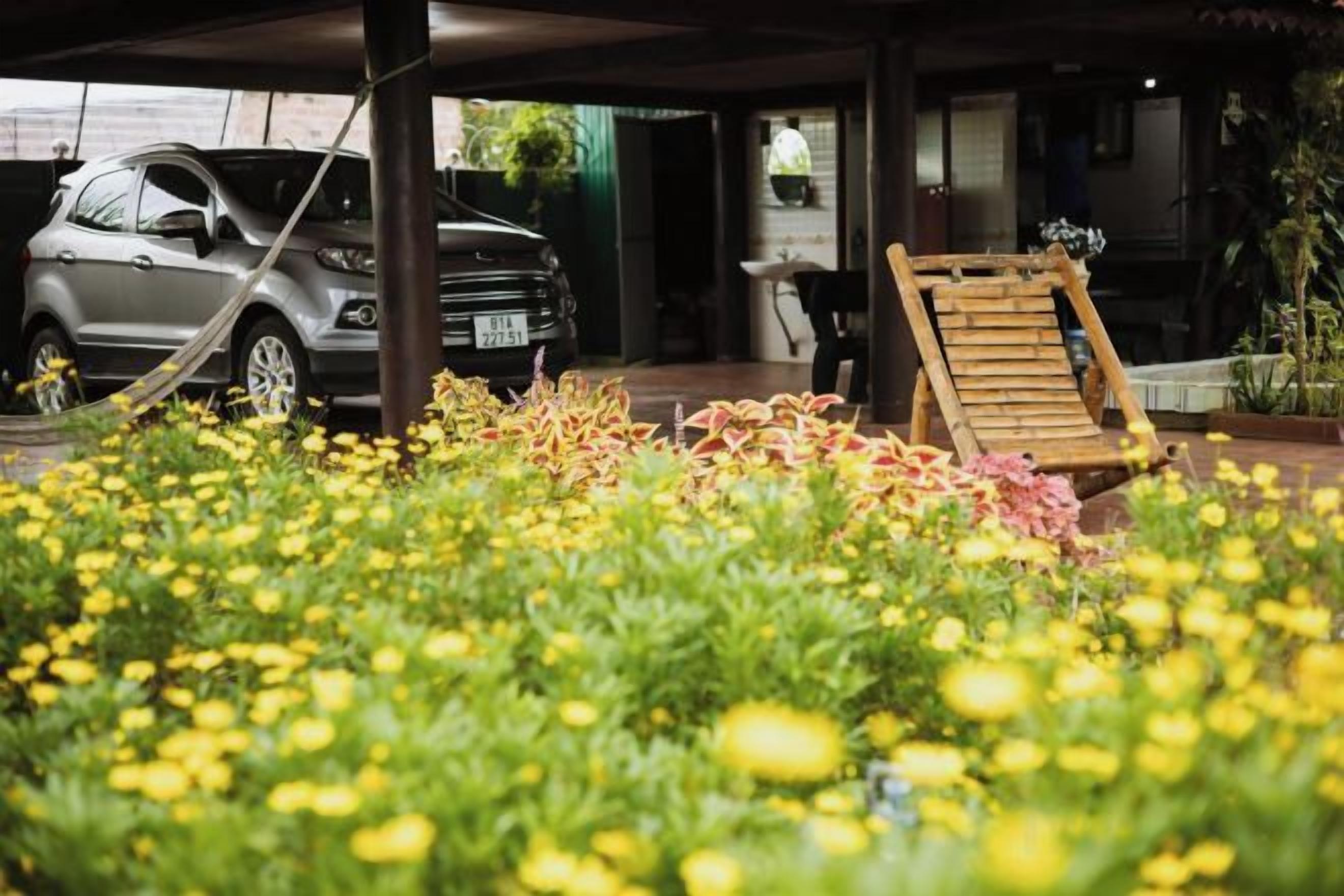 A car parked under a covered area with a chair and a table nearby, surrounded by a garden with yellow flowers.