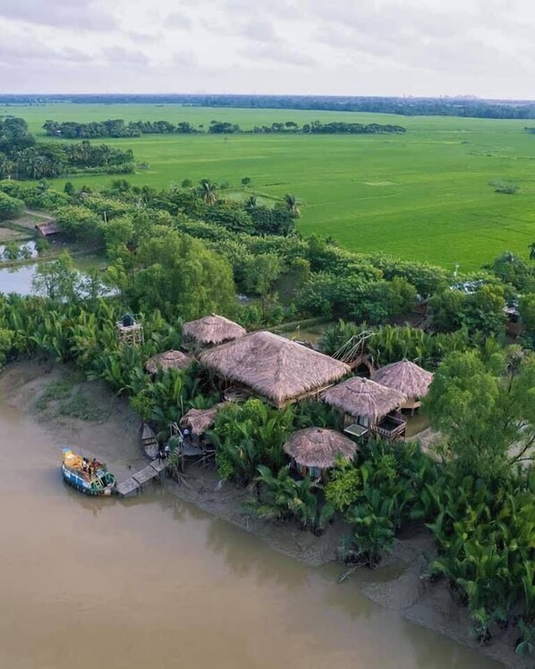 Alojamiento a orillas del río con cabañas de techo de paja, rodeado de exuberante vegetación y campos.
