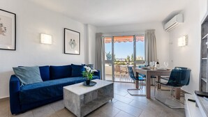 Un salon moderne avec un canapé bleu, une table basse et un coin repas comprenant une table et des chaises. Il y a un balcon avec vue sur la ville.