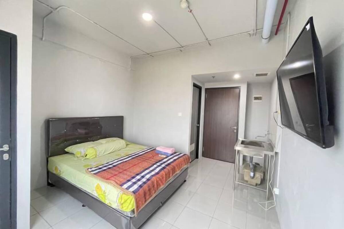 A bedroom with a bed, a TV, a small desk with a sink, and a door leading to another room.