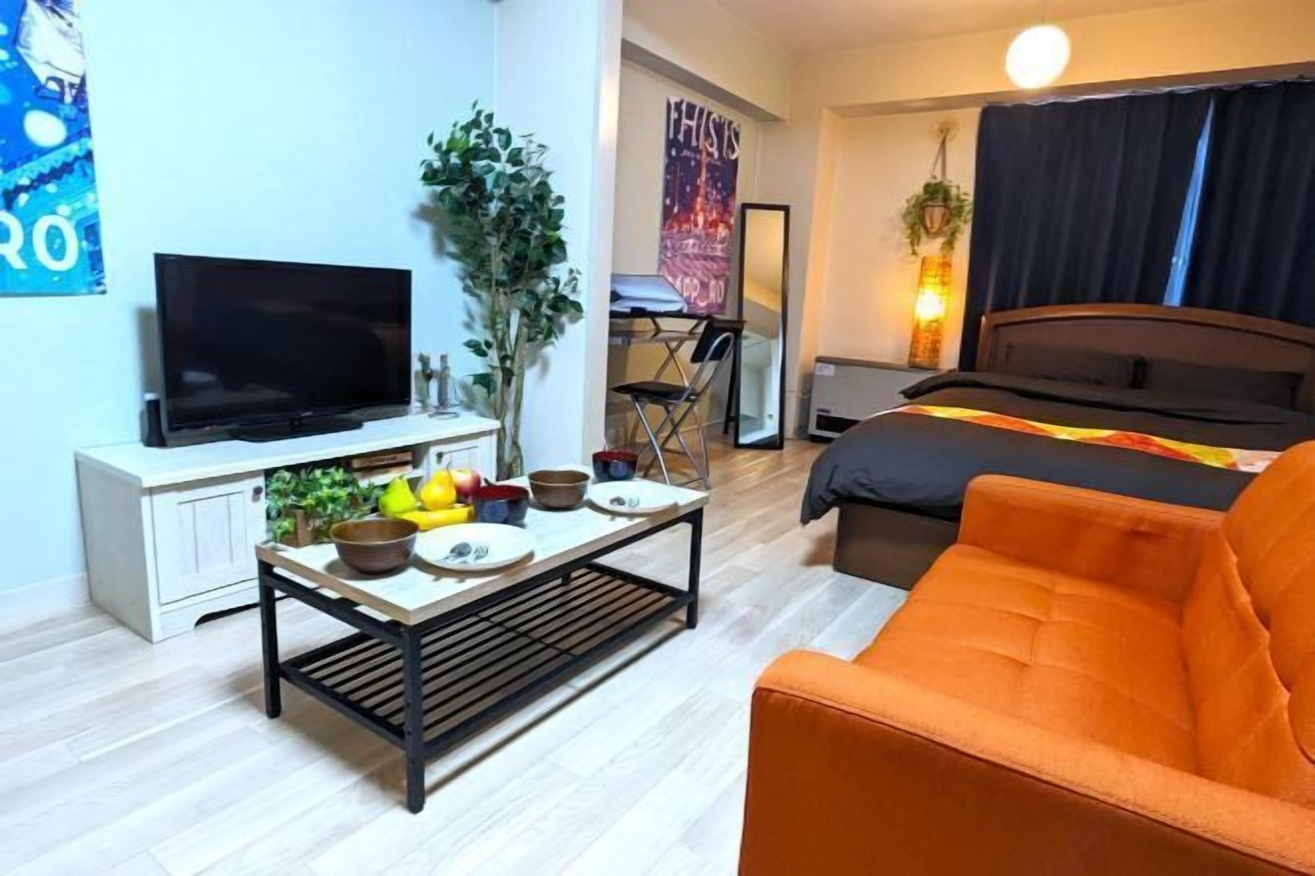 A compact hotel room with a bed, a TV, a small dining table with fruits, a sofa, and a mirror.