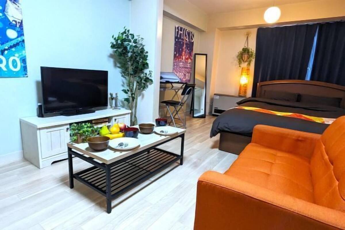 A compact hotel room with a bed, a TV, a small dining table with fruits, a sofa, and a mirror.