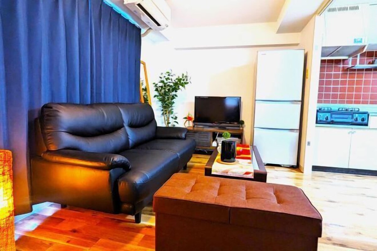A living room with a black sofa, a television, a small table with a pitcher, and a kitchen area with a refrigerator and stove.