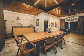 Un vaste espace de salle à manger avec une longue table en bois, de nombreuses chaises, des luminaires suspendus et, au fond, un coin cuisine.