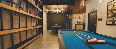A pool table with a blue felt surface, a wooden bookshelf, and a ceiling light fixture.