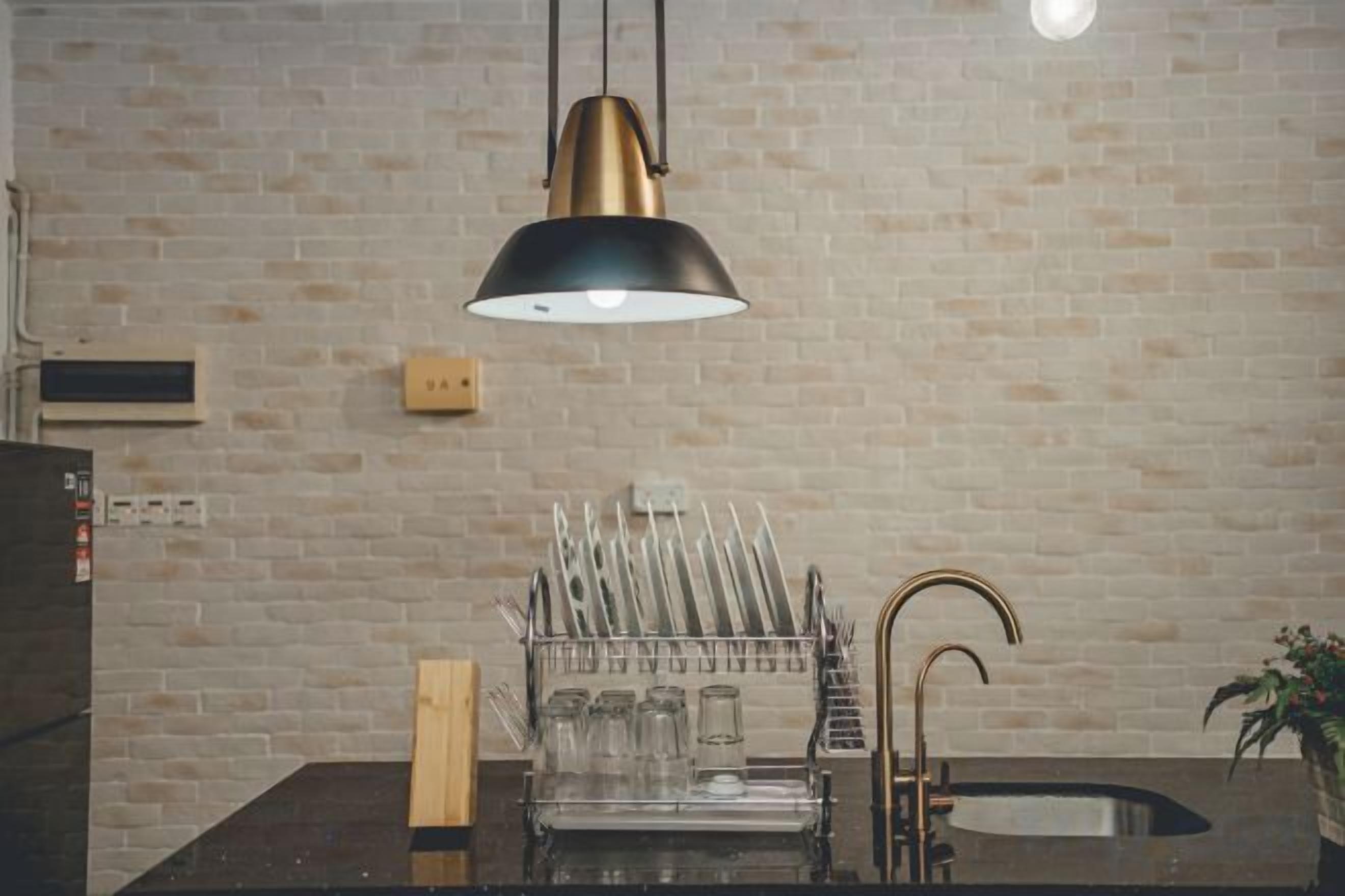 A kitchen with a brick wall, a black countertop, a sink, a faucet, a hanging light fixture, and a dish rack with utensils.