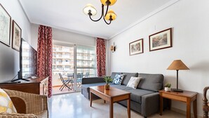 A living room with a sofa, coffee table, and a lamp. There is a balcony with red curtains and a view of buildings. - MalagaSuite Benalmadena Next to the Beach (Benalmádena)