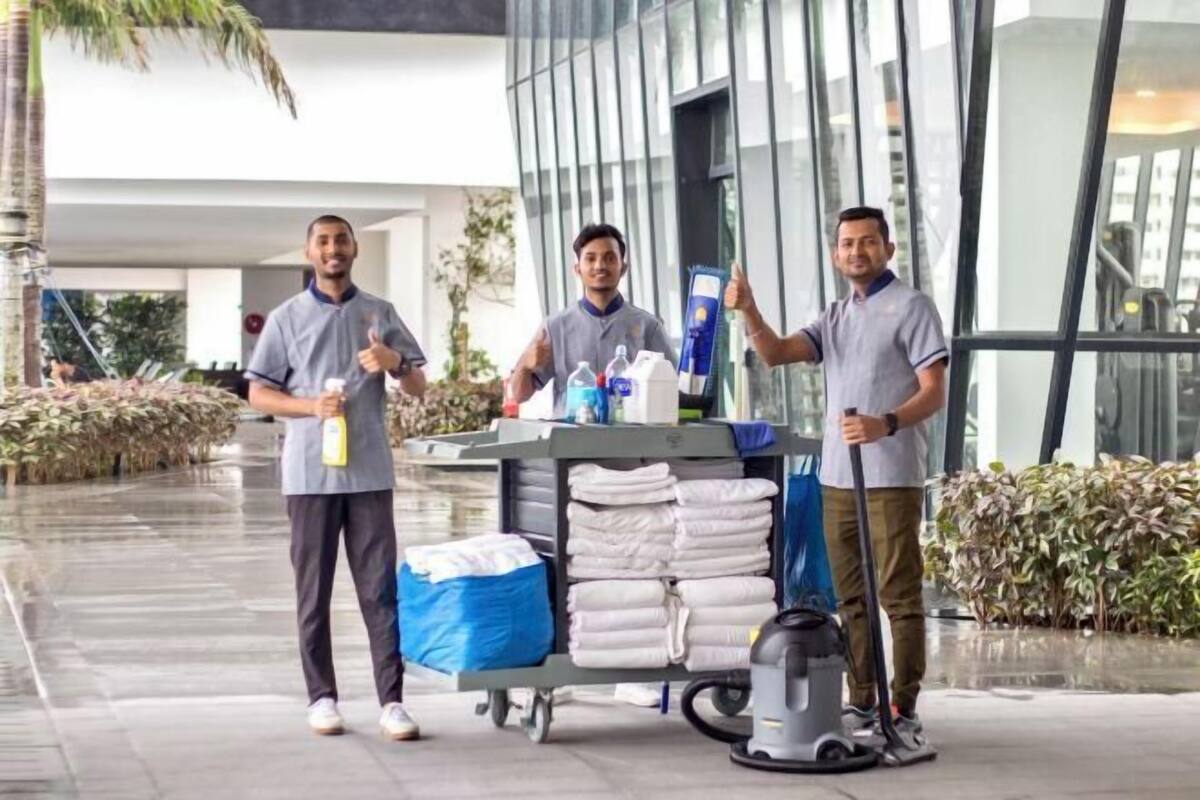 Trois personnes se tiennent devant un bâtiment, l’une d’entre elles tenant un chariot de nettoyage rempli de produits.