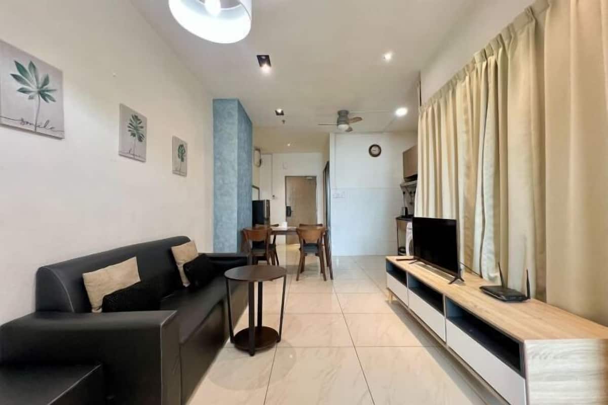 A modern living room with a sofa, a small table, a TV on a wooden stand, and a dining area in the background.