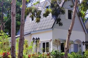 A two-story house with a sloped roof, a front porch, and a garden with various plants. - Pimbunya house (Phibun Mangsahan)