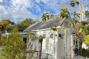 A white building with a sloped roof, a covered porch, and a small garden area with trees and a bench. - Pimbunya house (Phibun Mangsahan)