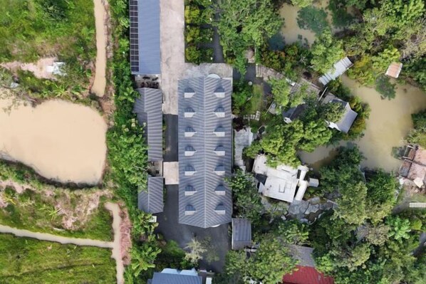 An aerial view of a residential area with a large house featuring a steeply pitched roof, surrounded by trees and a small pond.