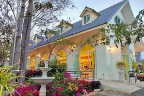 A two-story building with a covered porch, a fountain, and a garden with colorful flowers. - Pimbunya house (Phibun Mangsahan)