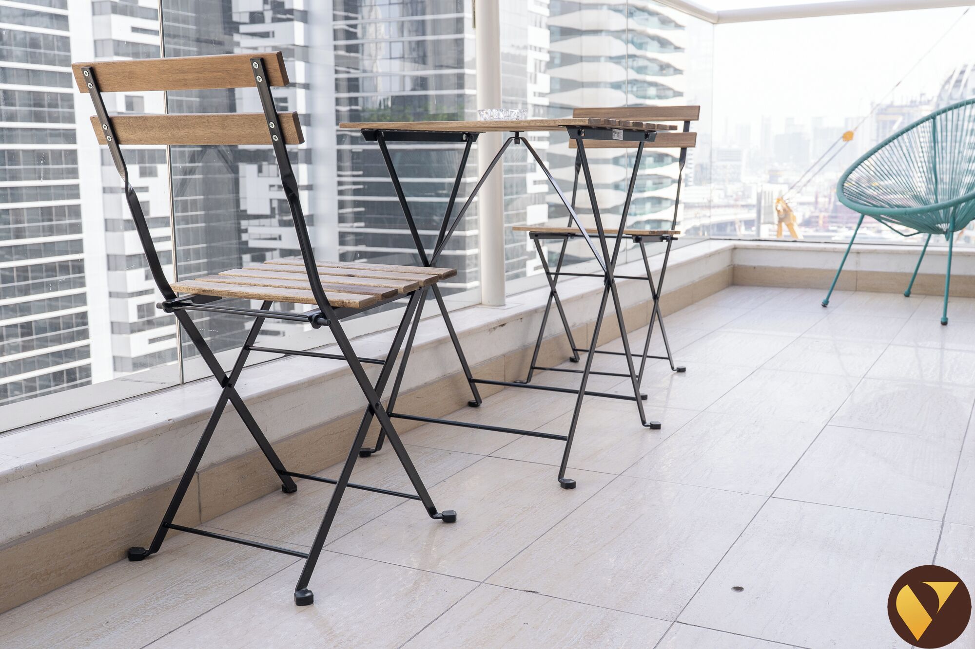A balcony with foldable chairs and tables, large windows with a city view, and a green chair in the background.