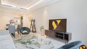A modern living room with a glass coffee table, a dark blue sofa, a wooden entertainment unit, and a large TV. - Vogue | Mon Reve near Dubai Mall | Burj Khailfa (Dubai)