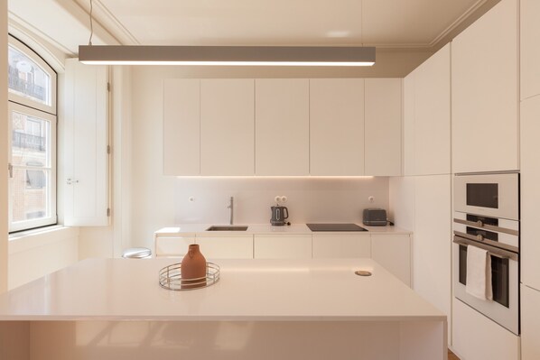 A modern kitchen with white cabinetry, a central island, a built-in oven, and a window providing natural light. - Nomad's Lisbon Call 2BDR Rua Augusta (Lisbon)