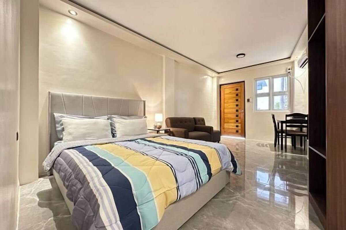 A modern bedroom with a large bed, a chair, a desk, and a wooden door.