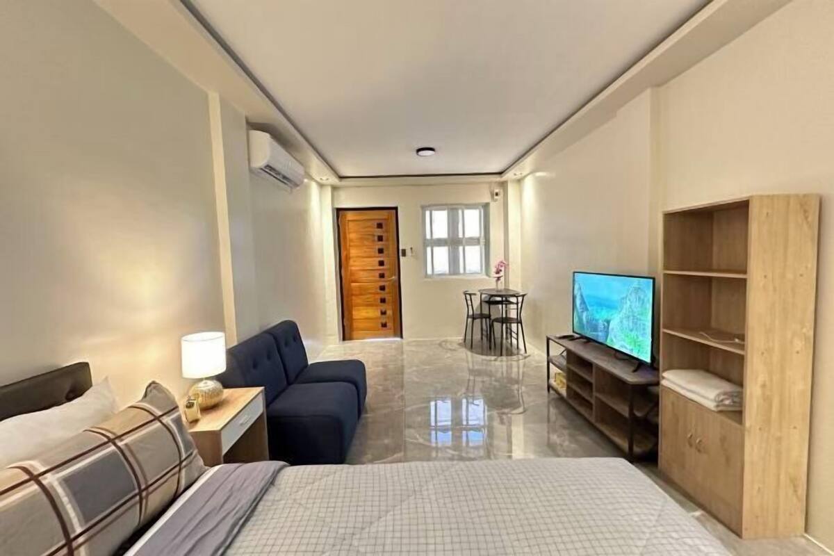 A hotel room with a bed, a desk with a lamp, a TV, a sofa, a dining table with chairs, and a wooden cabinet.