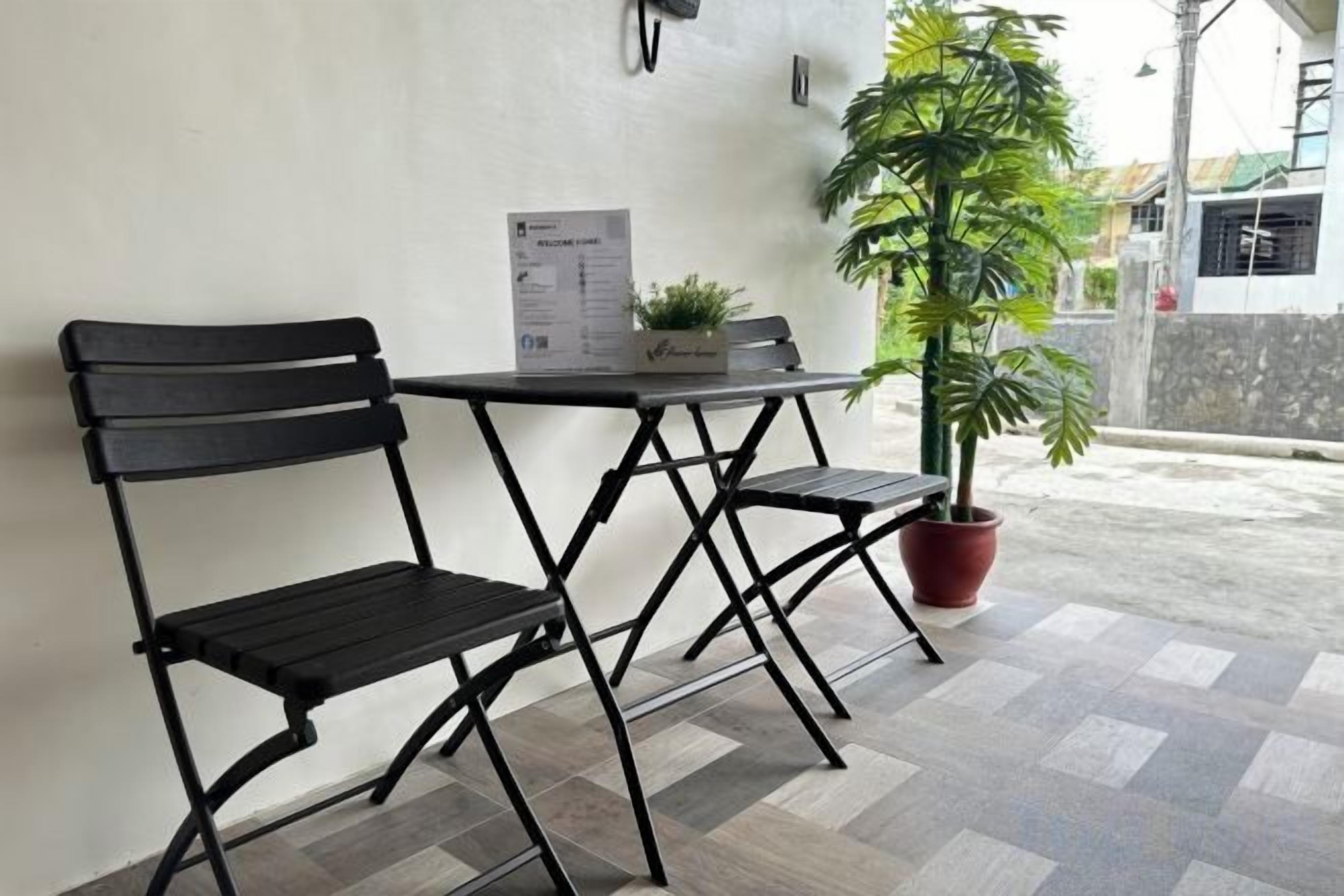 A patio area with two black metal chairs and a small table, a potted plant, and a menu stand.