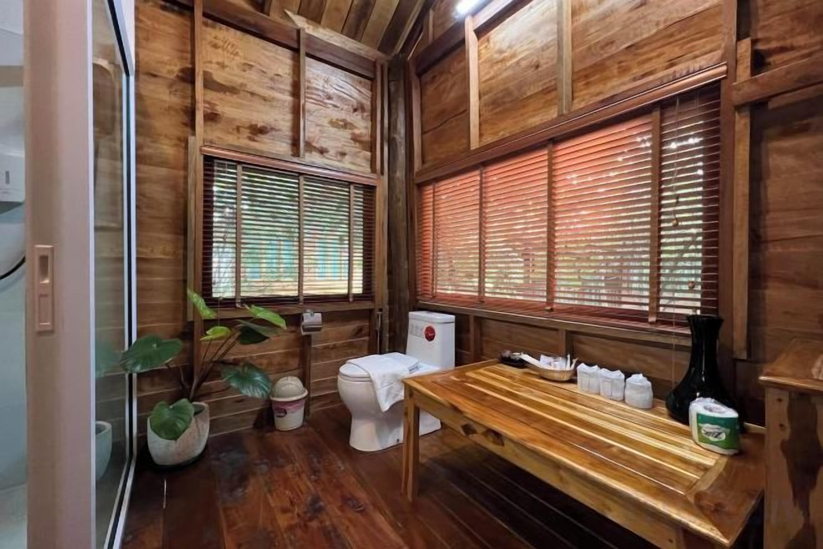 A wooden interior with a toilet, a sink, a wooden bench, and a window with blinds.