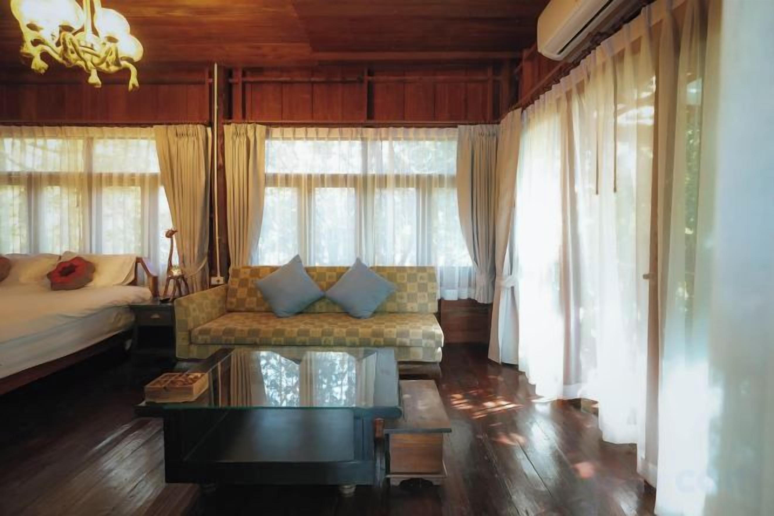 A room with a bed, a sofa with cushions, a glass coffee table, and a window with curtains.