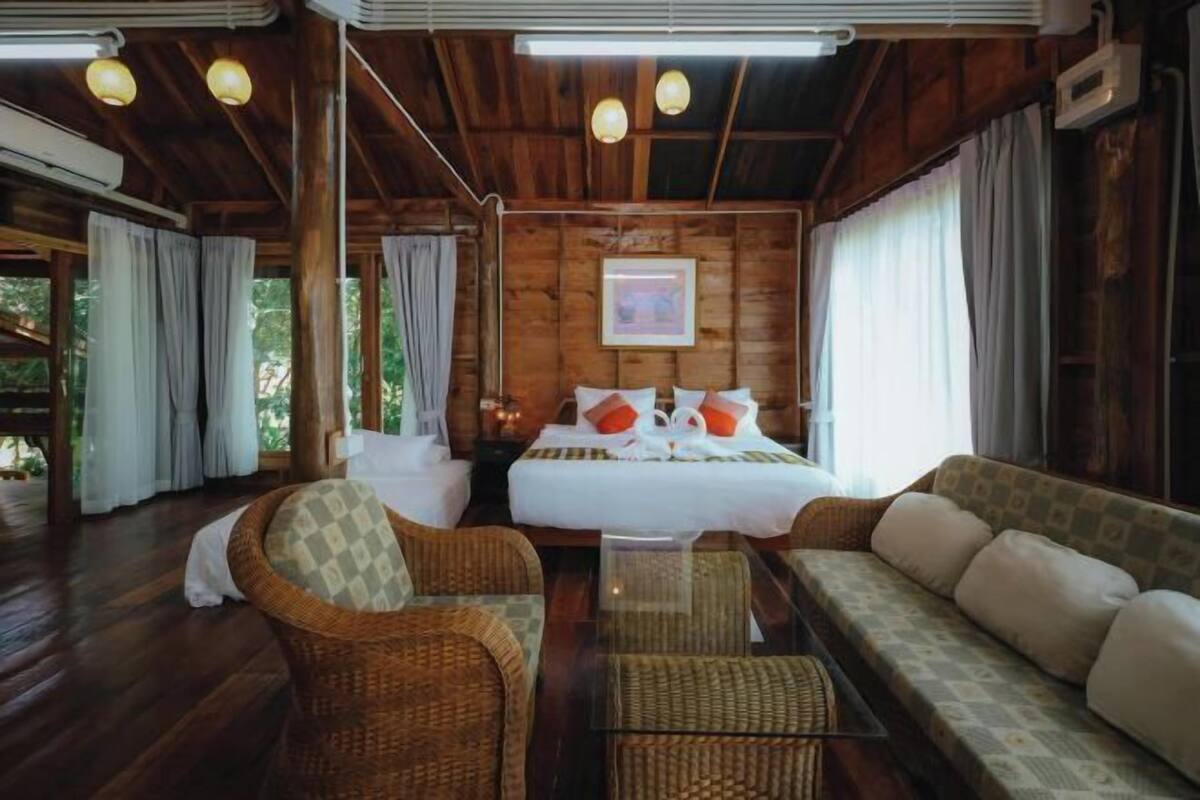 Een houten hutje met een bed, twee rieten stoelen, een bank en een glazen tafel.