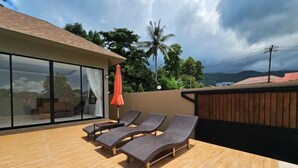 Une terrasse sur le toit avec trois chaises longues, un plancher en bois et un mur de verre donnant sur un paysage tropical.