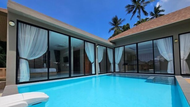 Un espace piscine moderne avec des parois en verre transparent et des rideaux blancs, entouré de palmiers et surplombé par un ciel dégagé.