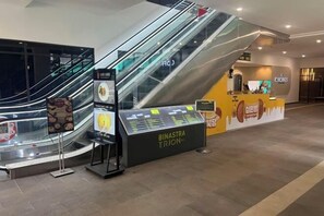 Espacio interior con una escalera mecánica, un mostrador con el rótulo 'BINASTRA TRION' y un cartel con imágenes de comida.