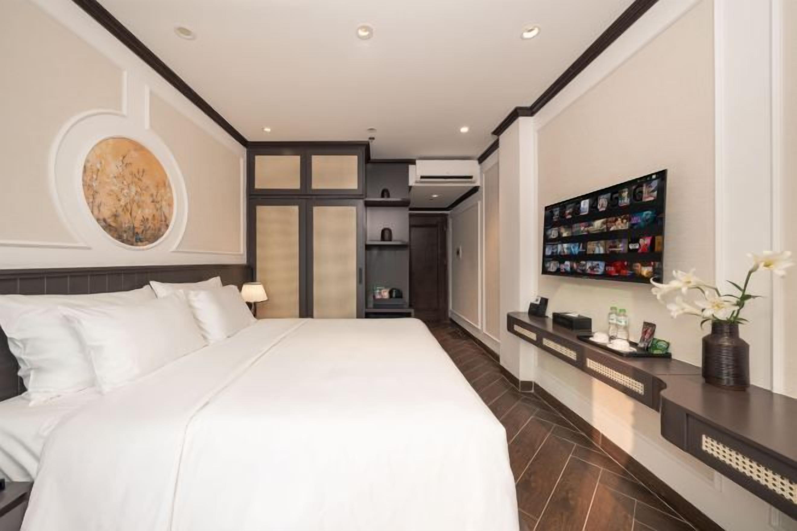 Une chambre d’hôtel avec un grand lit, un téléviseur fixé au mur et un comptoir de réception agrémenté d’un vase de fleurs.