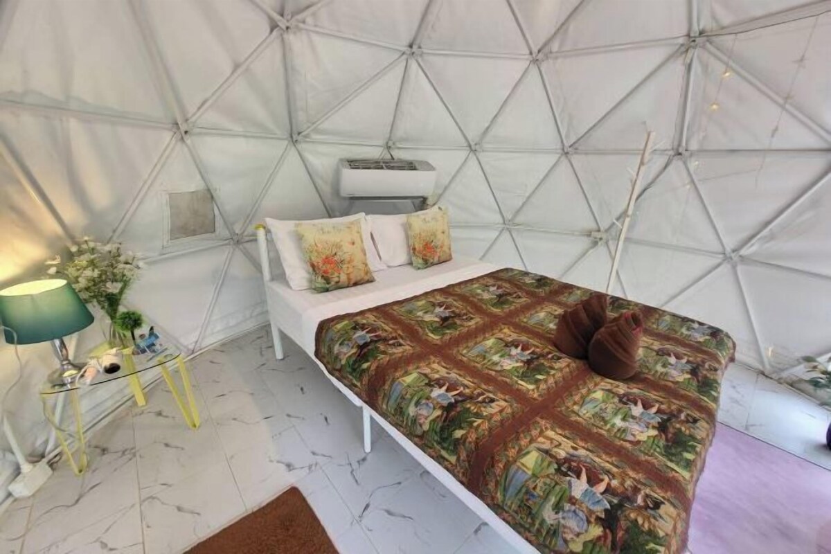Una habitación tipo tienda de campaña con una cama, una mesita con una lámpara y un equipo de aire acondicionado blanco.