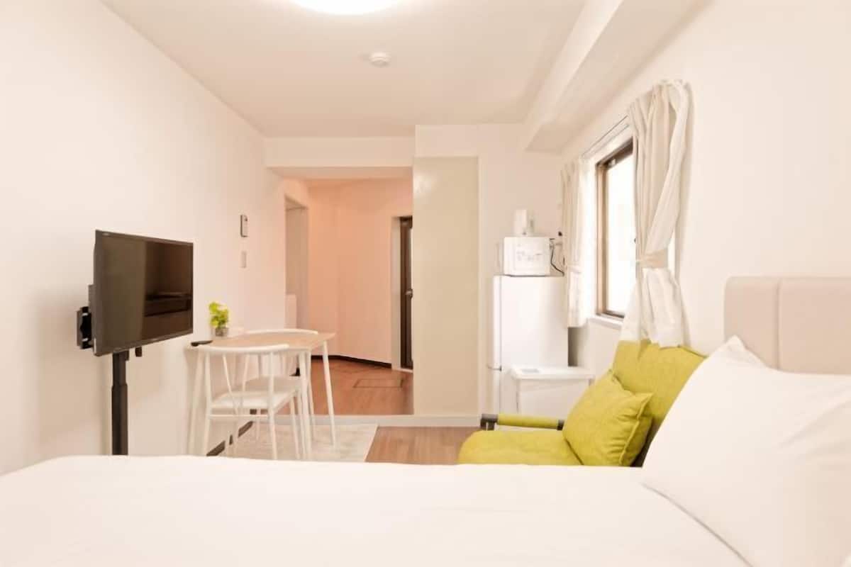 A bedroom with a bed, a TV, a small table with a chair, a window with curtains, and a door leading to another room.