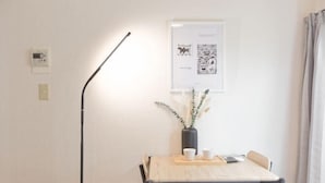 Una habitación minimalista con una mesa de madera, dos sillas, una lámpara de pie, un cuadro enmarco y un florero con plantas.