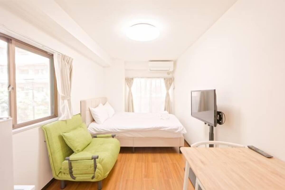 A small, bright room with a bed, a green chair, a desk, and a TV.