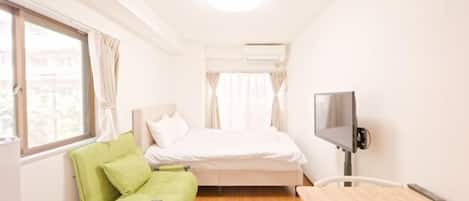 A small, bright room with a bed, a green chair, a desk, and a TV.