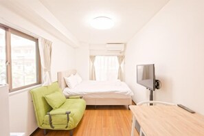 A small, bright room with a bed, a green chair, a desk, and a TV.